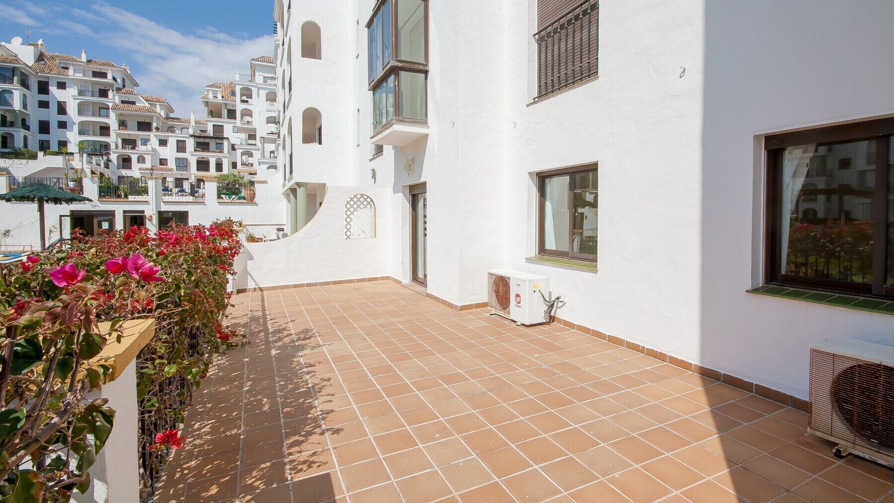 Un bâtiment blanc doté d’un patio carrelé, d’un petit climatiseur et d’une jardinière ornée de fleurs roses.