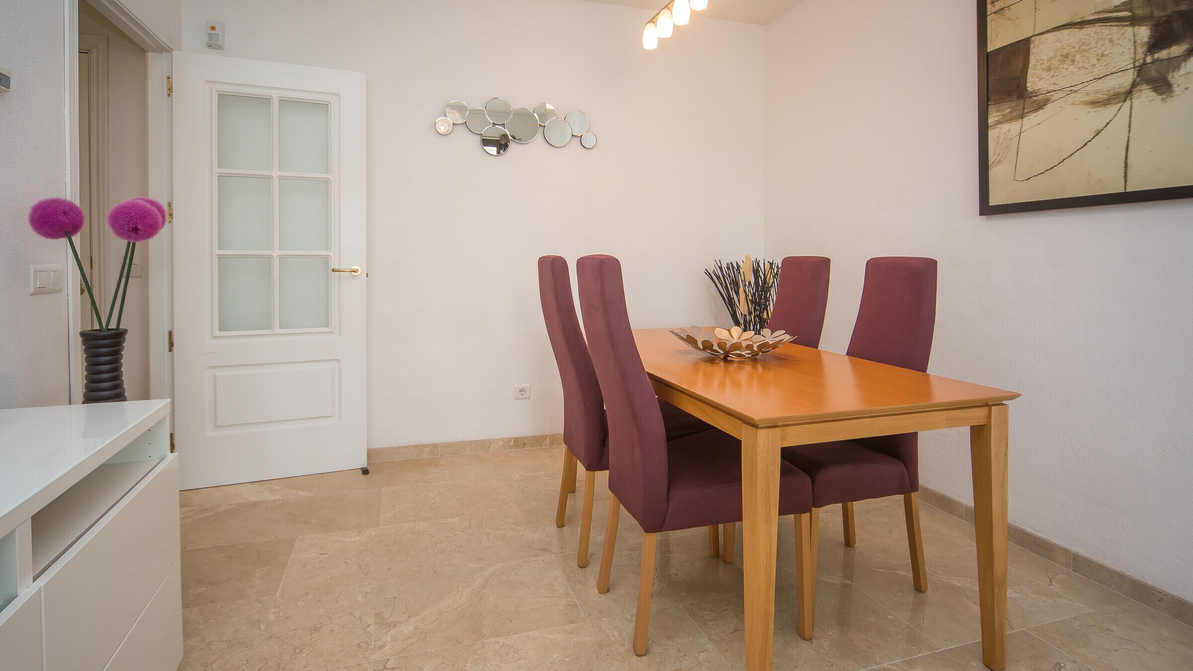 Een eetgedeelte met een houten tafel en vier paarse stoelen, een witte deur en een spiegel aan de muur.