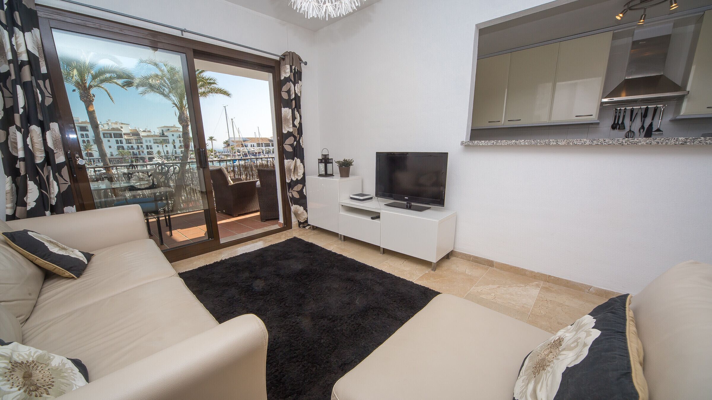 A living room with a sofa, a TV, a small white cabinet, and a balcony with a view of buildings and palm trees.