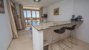 Una cocina con encimera de granito, dos taburetes de bar, un televisor y vista al mar a través de amplias ventanas.