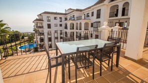Varanda com mesa e cadeiras de vidro, com vista para uma piscina e um edifício branco com várias varandas.
