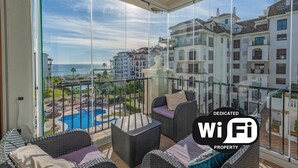 En balkon med fletmøbler, udsigt til en pool og en by silhuet i baggrunden.