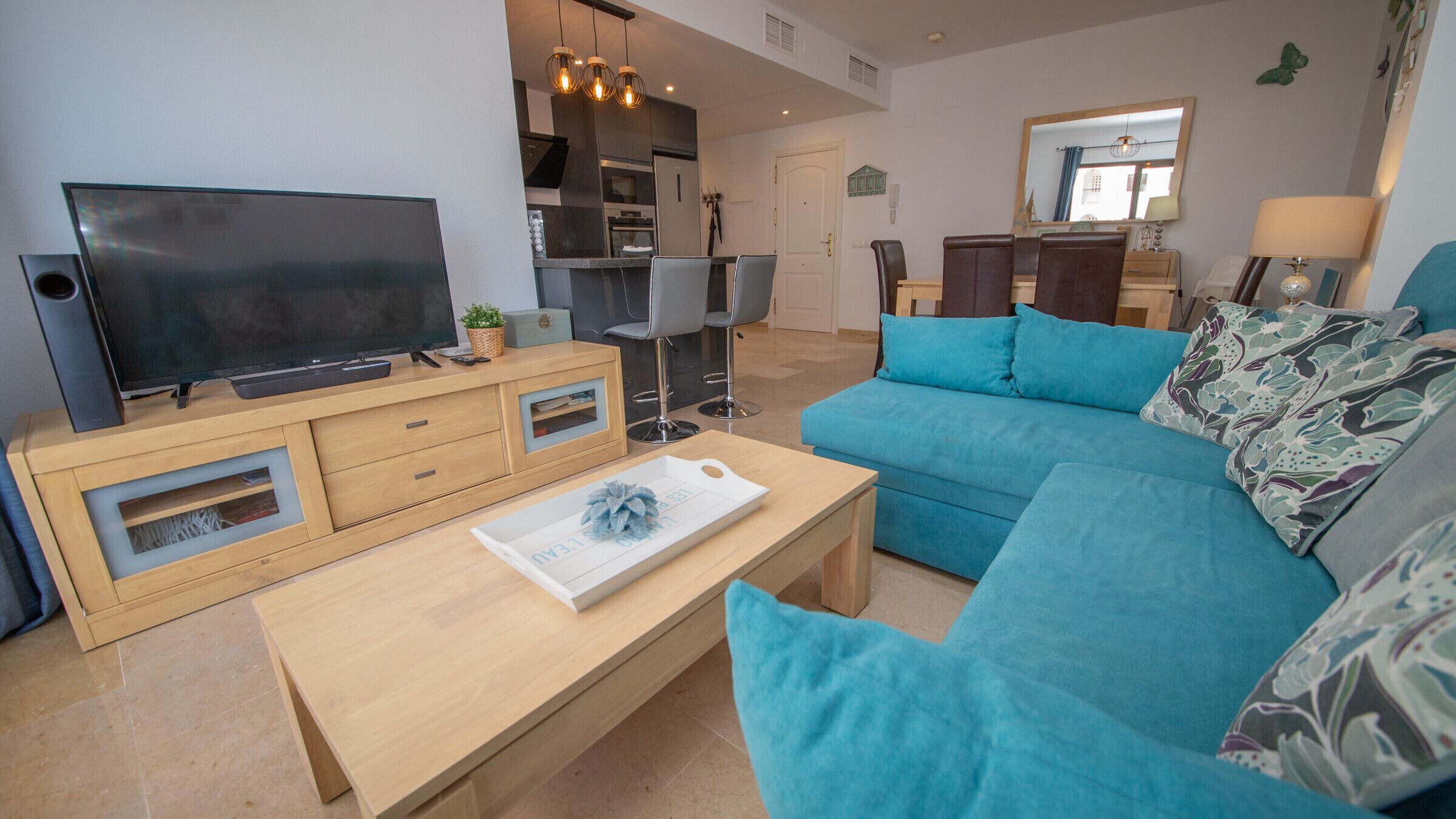 Een moderne woonkamer met een grote flatscreen televisie, een houten salontafel, een blauwe bank en een keuken in de achtergrond.