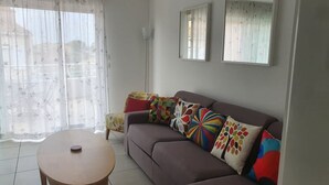 A living room with a brown sofa, colorful pillows, a small wooden table, and a large window with sheer curtains. - Sunny apartment overlooking Etang de Thau (5 km from the sea) (marseillan)