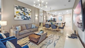 A modern living room with a sofa, coffee table, and artwork. There is a dining area with chairs and a table, and a kitchen in the background.