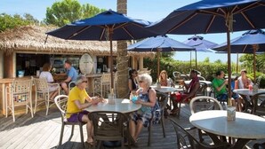 Un espace de restauration en plein air avec des tables rondes et des parasols bleus, entouré d’un bar à toit de chaume.