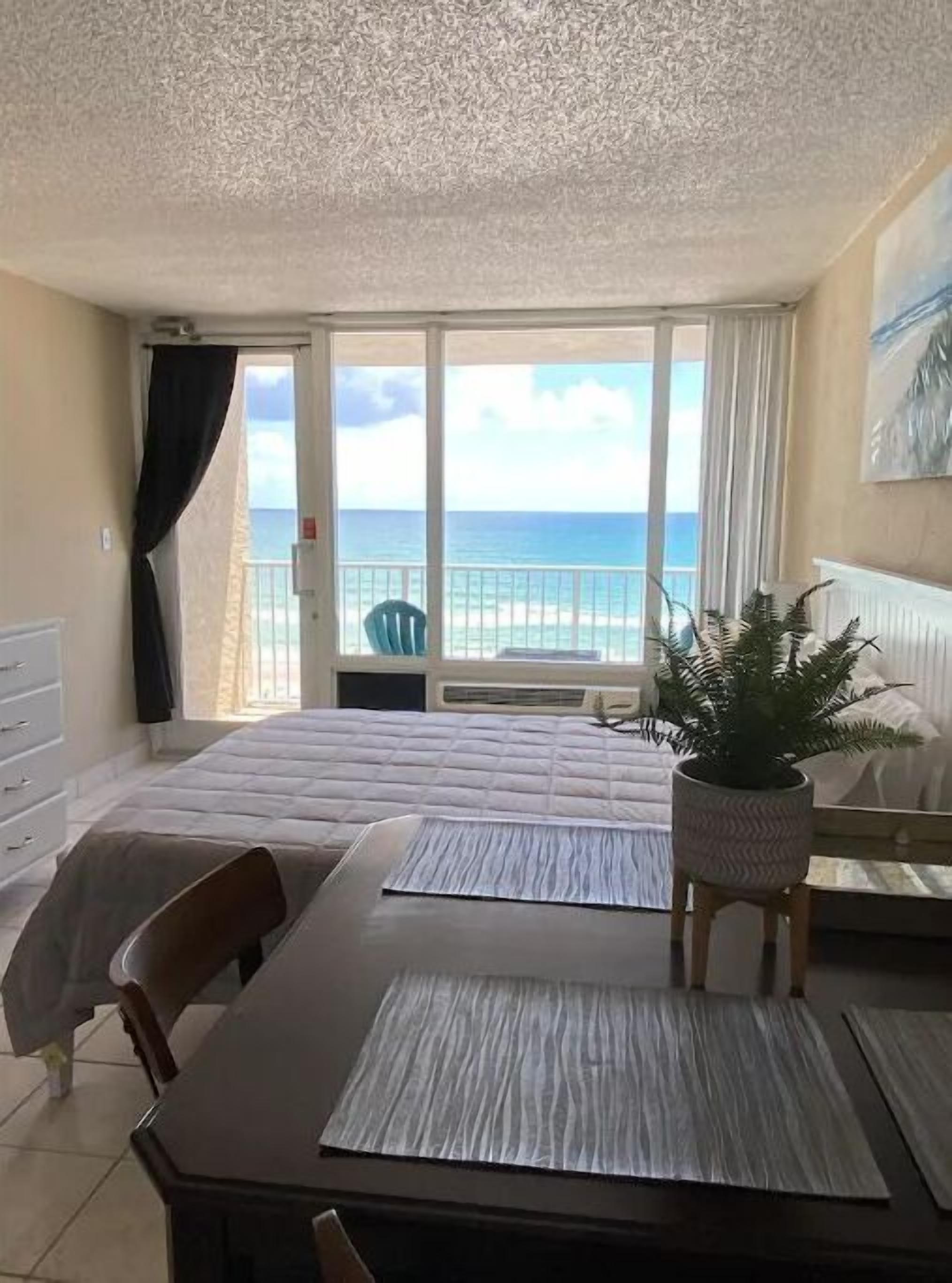 A bedroom with a bed, a dining table, chairs, a potted plant, and a view of the ocean through large windows.