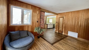 A modern interior with wooden paneling, a grey sofa, a potted plant, and a kitchen in the background. - 2355/2 · Villa des pins (Saint-Gabriel-de-Valcartier)