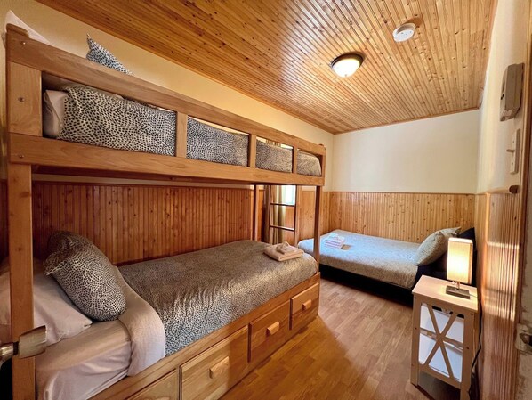 A wooden bunk bed room with a single bed on the lower bunk and a single bed on the upper bunk. There is a small bedside table with a lamp on the right side.