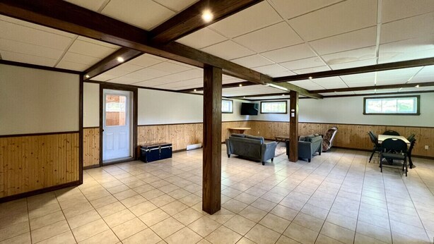 A spacious room with a high ceiling, wooden beams, tiled flooring, a seating area with sofas and a table, and a television mounted on the wall. - Cottage Montagne - Vallée Jeunesse site (Saint-Gabriel-de-Valcartier)