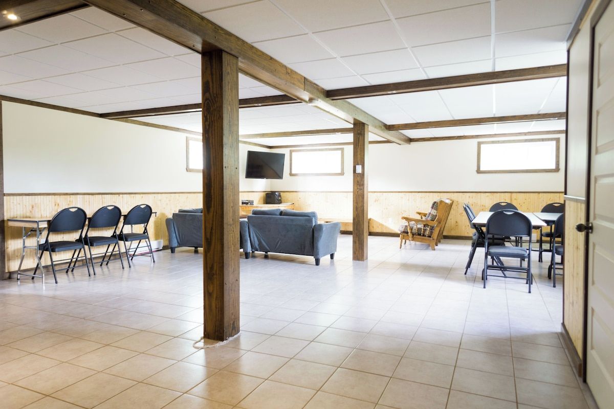 A spacious room with a high ceiling, wooden beams, and a tiled floor. There are several tables and chairs arranged for dining or meetings.