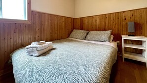 A bedroom with a wooden ceiling, a bed with patterned bedding, a window, and a bedside table with a lamp. - Cottage Montagne - Vallée Jeunesse site (Saint-Gabriel-de-Valcartier)