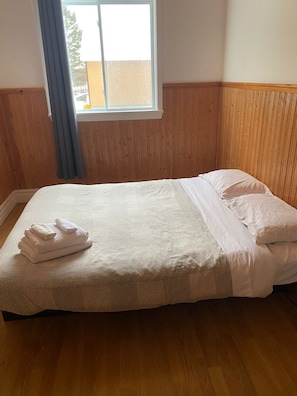 Un lit simple avec une couette et des oreillers blancs, une fenêtre donnant sur un bâtiment à l’extérieur, et des lambris en bois sur les murs.