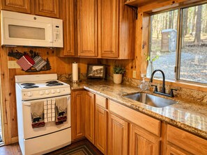 A kitchen with wooden cabinets, a white stove, a microwave, a sink, and a window with a view of trees. - Weekend Getaway Relax & Recharge 5 acres Nature Retreat & Dog Heaven!  (Mokelumne Hill)