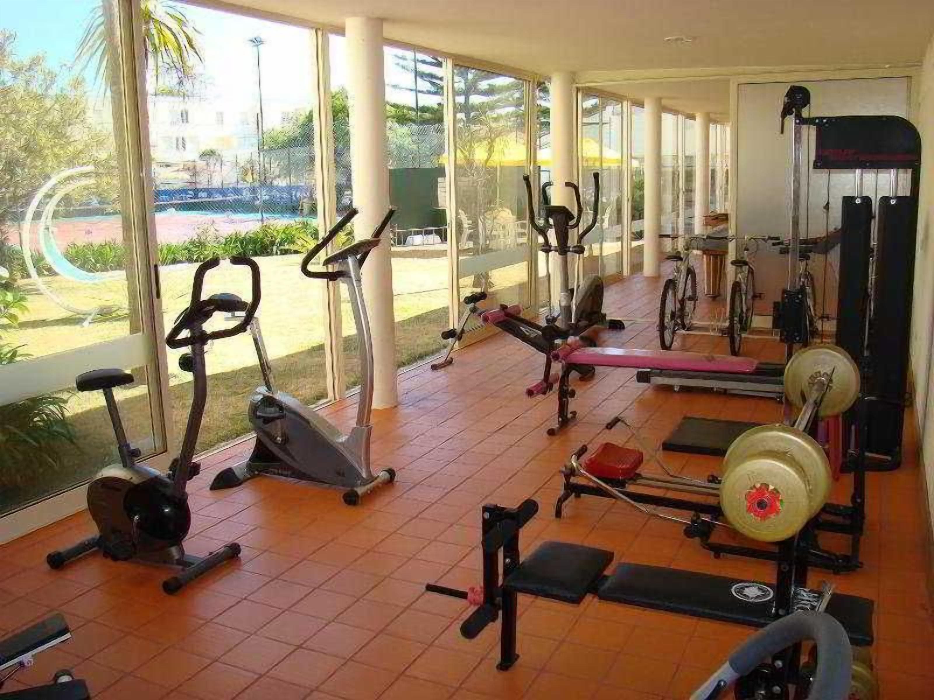 Ein Fitnessstudio mit unterschiedlichen Trainingsgeräten, darunter Laufbänder, Ergometer und Kraftgeräte, mit großen Fenstern, die Ausblicke ins Freie bieten.