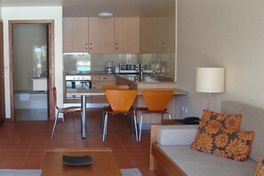 A compact living space with a kitchenette, a dining area with two orange chairs, a sofa with floral cushions, and a lamp. - Clube Pinhal Da Foz (Esposende)