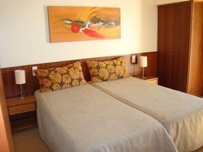 A double bed with a neutral-colored bedspread and two decorative pillows, a wooden headboard, a nightstand with a lamp, and a wooden wardrobe. - Clube Pinhal Da Foz (Esposende)