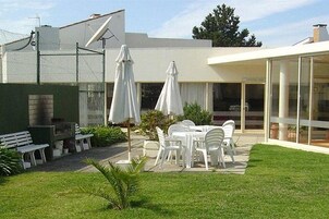 Un patio con sillas y sombrillas de plástico blanco, un pequeño jardín y un edificio con ventanas amplias y una zona techada.
