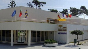 A building with multiple flags, a sign reading "CLUBE DE FORA," and a small garden area. - Clube Pinhal Da Foz (Esposende)