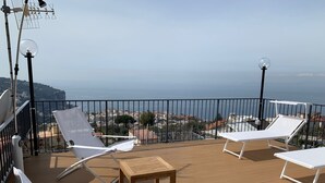 A wooden deck with white lounge chairs and a table, overlooking a coastal cityscape. - Casa Paola 2 (Vico Equense)