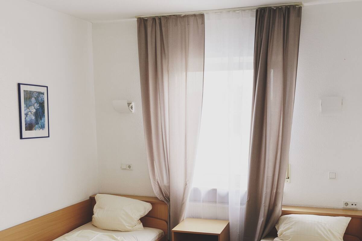 Una habitación pequeña y limpia con dos camas individuales, una mesa de madera y un ventanal con cortinas traslúcidas.