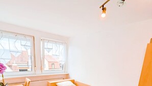 Una habitación pequeña y luminosa con una cama de madera, una mesa y un ventanal con vistas a edificios.