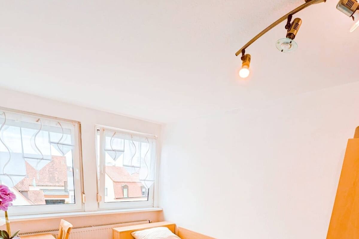 Una habitación pequeña y luminosa con una cama de madera, una mesa y un ventanal con vistas a edificios.