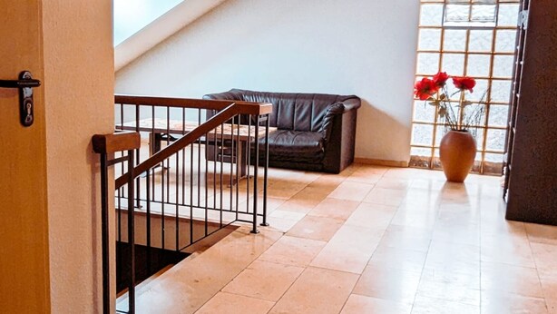 Un pasillo amplio con una escalera que sube a una zona de altillo, un sofá de cuero y un jarrón con flores.