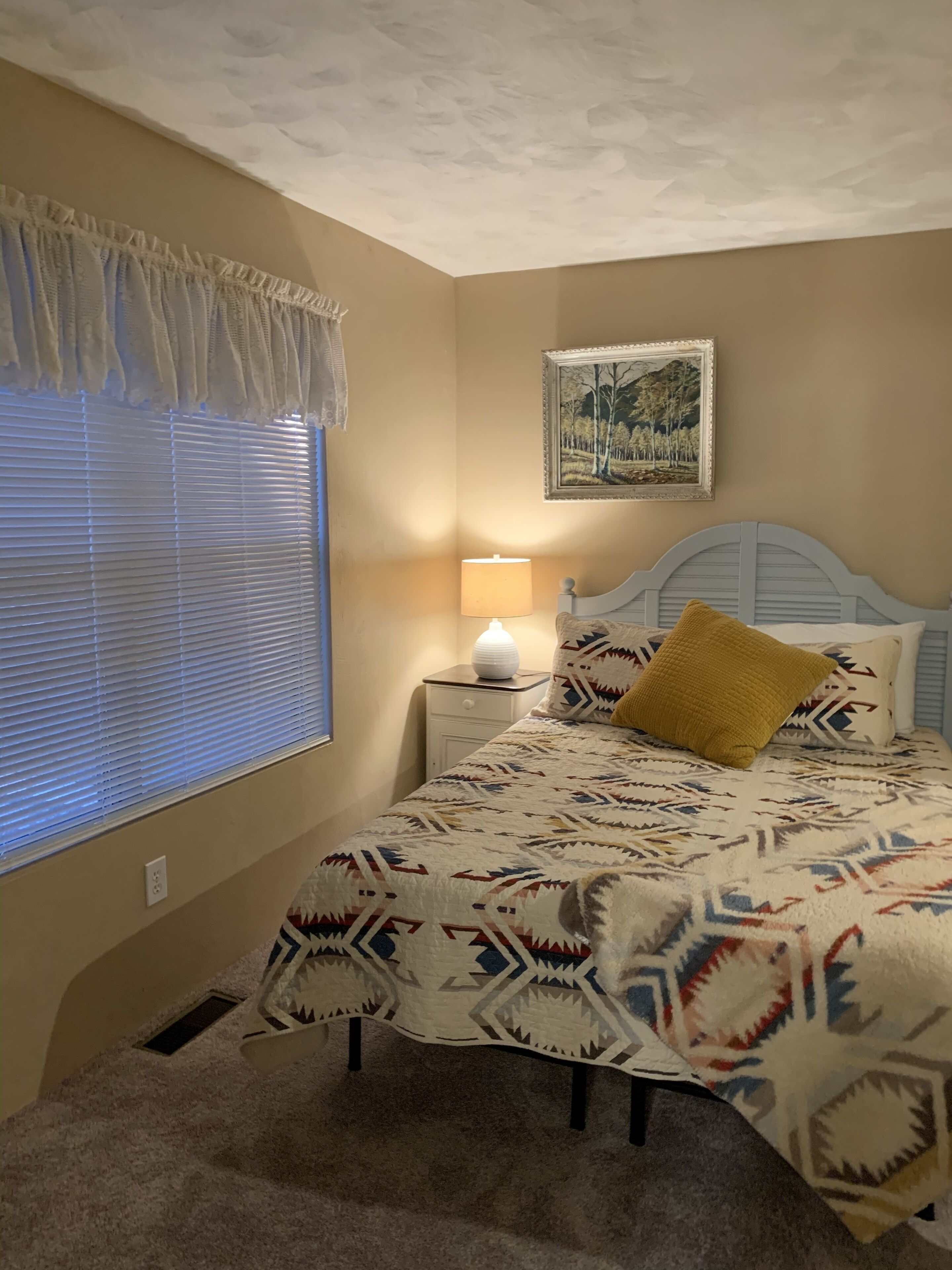 A bedroom with a bed, a nightstand, a lamp, a window with blinds, and a wall-mounted picture.