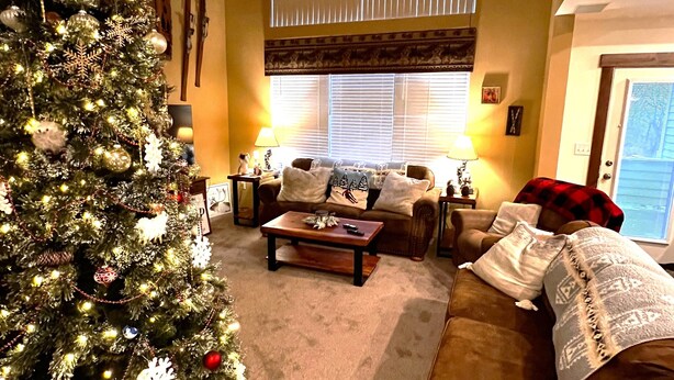 A cozy living room with a large Christmas tree, a brown sofa, a coffee table, and a window with blinds. - Charming Rustic Townhome in the Heart of the Villages at Seven Springs (Champion)