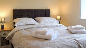 Un lit bien fait, avec du linge de lit blanc et deux oreillers, une table de chevet avec une lampe, et une fenêtre laissant entrer la lumière du jour.