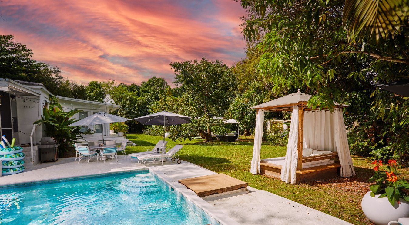 Une piscine avec une terrasse en bois, des chaises longues et des parasols. On aperçoit un lit à baldaquin et une maison au fond.
