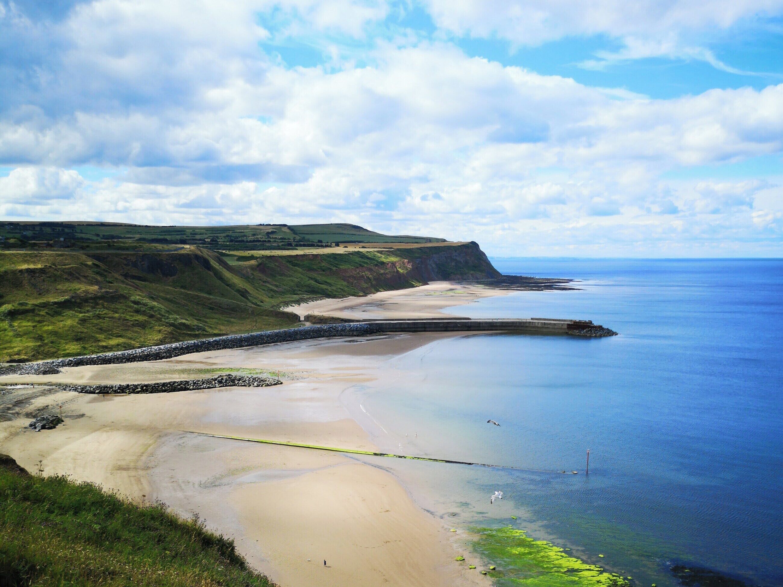 A coastal landscape with a sandy beach, a rocky breakwater, and a view of the sea.