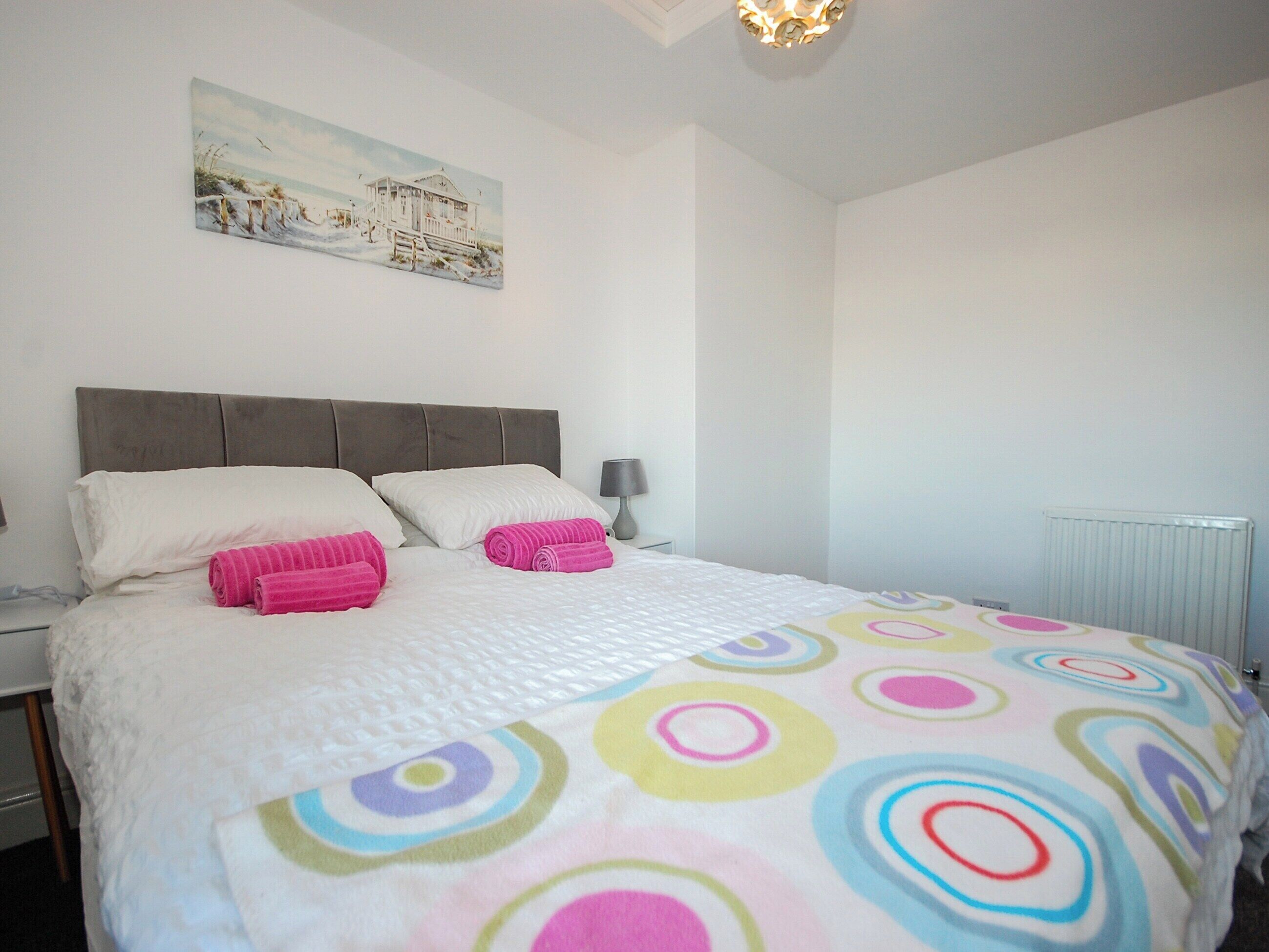 Un lit bien fait, recouvert d’une couvre-lit aux motifs circulaires et colorés, avec une tête de lit grise et un tableau représentant une scène de plage accroché au mur.