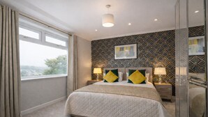 A hotel room with a large bed, two bedside tables with lamps, a window with curtains, and a patterned wall. - HC Property - Taylor Avenue (Durham)