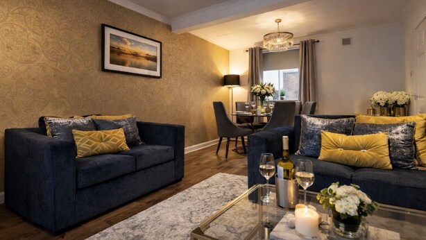 A modern living room with dark blue sofas, gold and black cushions, a dining area with a chandelier, and a view of the city through large windows. - HC Property - Taylor Avenue (Durham)