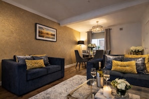 A modern living room with dark blue sofas, gold and black cushions, a dining area with a chandelier, and a view of the city through large windows. - HC Property - Taylor Avenue (Durham)