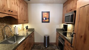 Cocina compacta con encimeras de granito, armarios de madera, pileta de acero inoxidable, microondas y basurero.