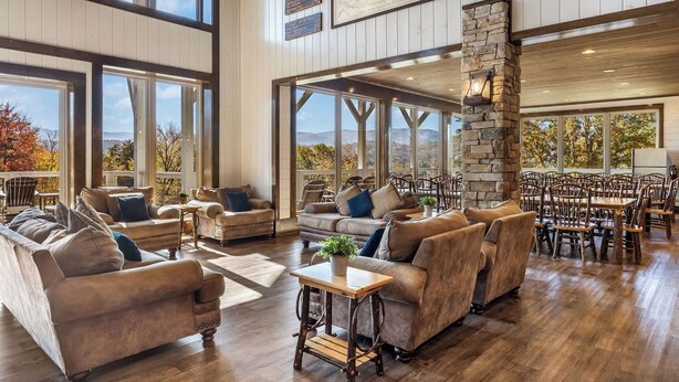 A spacious indoor area with multiple seating arrangements, large windows offering views of the outdoors, and a stone pillar. - Grand LeConte Lodge | 21BR Luxury Estate w/ Pool, Theater, Arcade, Sleeps 96 (Pigeon Forge)