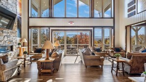 A spacious, well-lit lounge area with multiple seating options, large windows offering scenic views, and a stone accent wall. - Grand LeConte Lodge | 21BR Luxury Estate w/ Pool, Theater, Arcade, Sleeps 96 (Pigeon Forge)