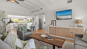 Een ruime woonkamer met een bank, salontafel en een aan de muur bevestigde televisie. Achtergrond zichtbaar is een keuken, en op de muur staat een bordje met de tekst 'BEACH HOUSE'.