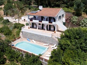 A two-story house with a swimming pool, surrounded by greenery and a stone wall.