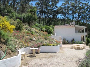 A white villa with a balcony, surrounded by a garden with various plants and flowers.