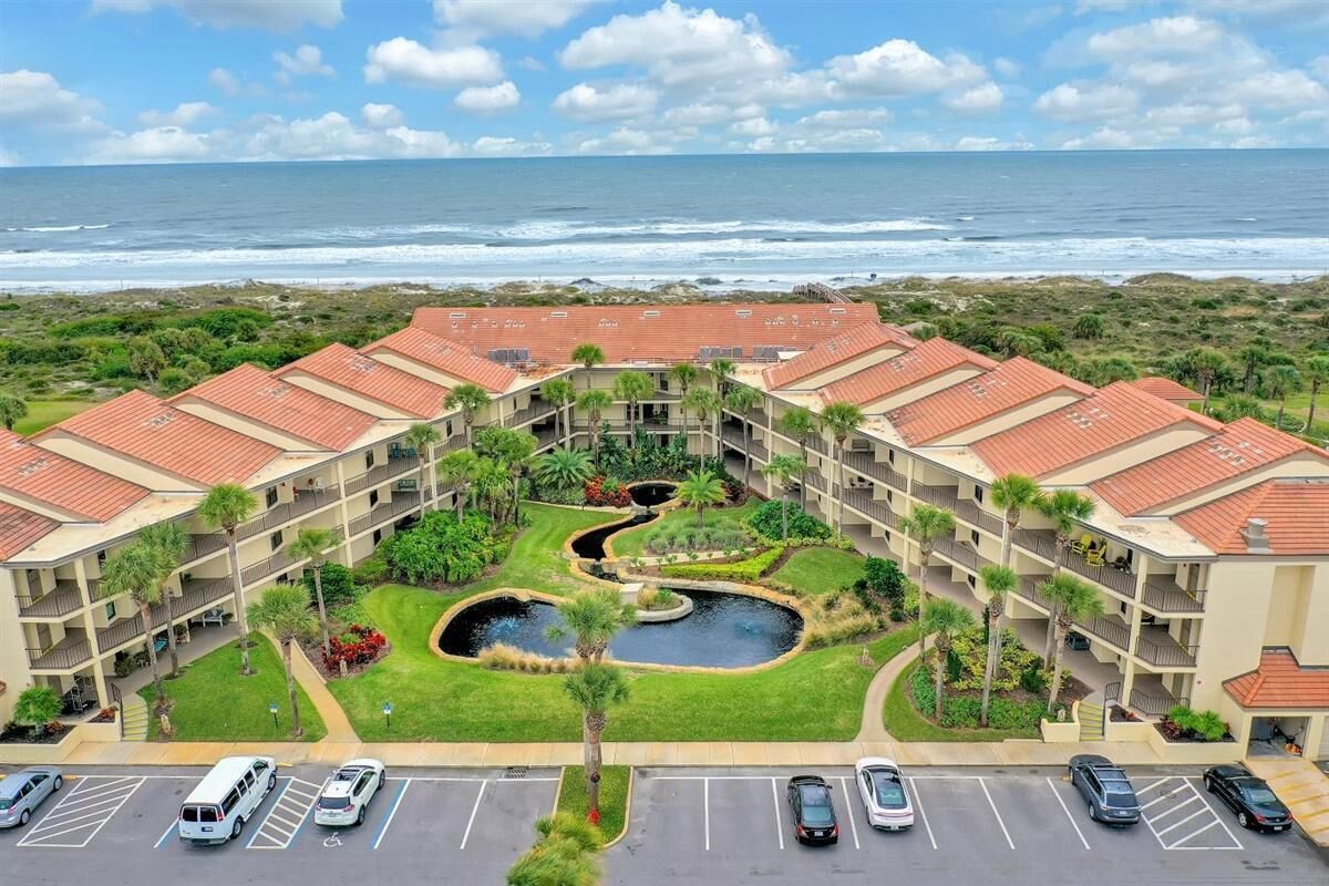 A hotel with a central courtyard featuring a pond, surrounded by lush greenery and palm trees. The building has a red-tiled roof and multiple balconies.