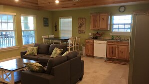 A living room with a dark brown sofa, a coffee table, and patterned cushions. There is a dining table with chairs, a kitchen area with wooden cabinets, a refrigerator, and a clock on the wall. - 10% Discount- Hackberry Hideaway II Water View On Lake Calcasieu (Hackberry)