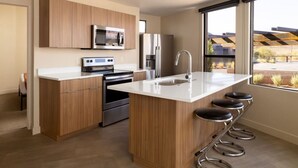 A modern kitchen with wooden cabinets, stainless steel appliances, a central island with stools, and large windows offering a view.
