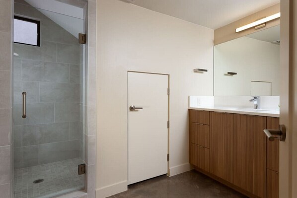 A modern bathroom with a glass shower enclosure, a white door, wooden cabinets, and a mirror above the sink. - Modern Desert Dwelling W/ Garage (Ivins)