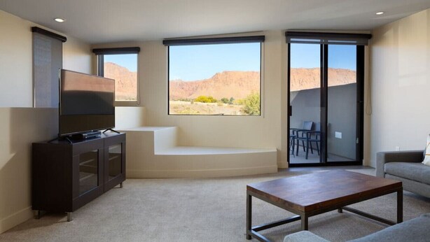 A modern living room with a flat-screen TV, a sofa, a coffee table, and a large window offering a view of a desert landscape.