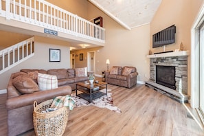 A spacious living room with a large brown leather sectional sofa, a coffee table, and a fireplace. A staircase leads to a balcony with a white railing.