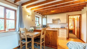 Une cuisine chaleureuse avec des poutres en bois, un coin repas avec une table et des chaises, et une vue sur l’extérieur par une fenêtre.